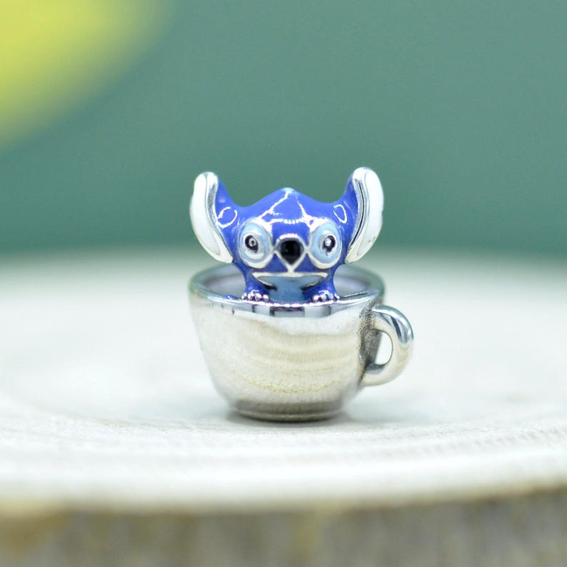 May include: A silver-toned metal charm featuring a blue enamel character with large ears and dark eyes, positioned inside a small silver-toned cup. The character has white accents on the ears and a smooth, polished finish on the cup exterior.