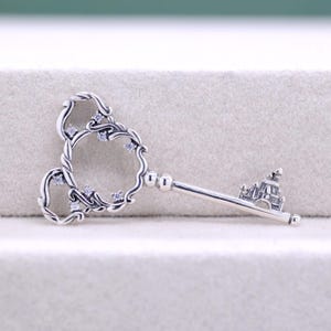 May include: A silver-colored key pendant with a decorative Mickey Mouse-shaped top, adorned with small, clear gemstones. The key's stem features a miniature castle design, adding a whimsical touch to this jewelry piece.
