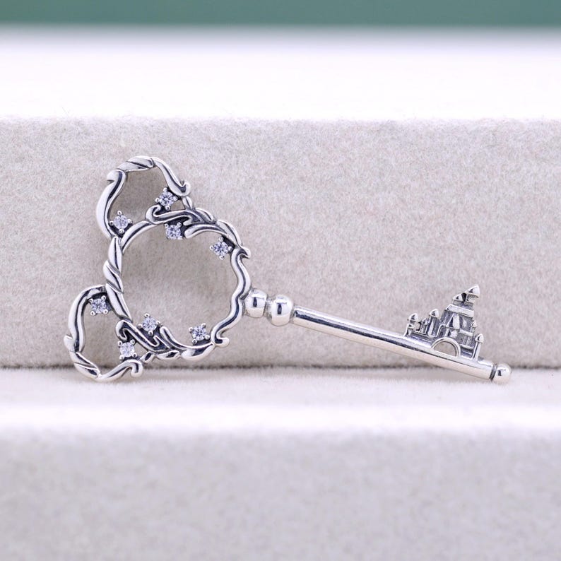 May include: A silver-colored key pendant with a decorative Mickey Mouse-shaped top, adorned with small, clear gemstones. The key's stem features a miniature castle design, adding a whimsical touch to this jewelry piece.