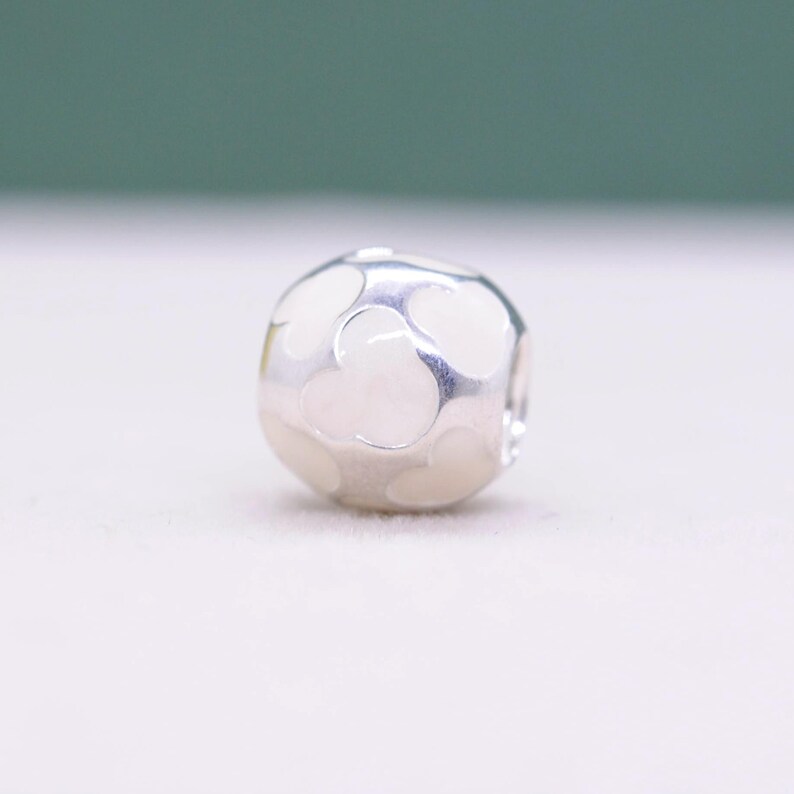 May include: A silver charm bead with a rounded, geometric shape. The bead features a pattern of white heart-shaped cutouts. The charm is designed for bracelets and necklaces, offering a decorative element.
