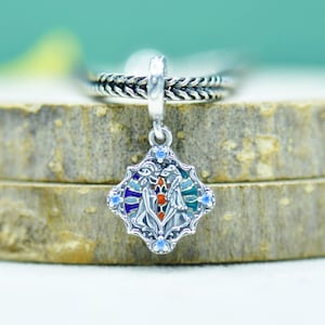 Pode incluir: Um pingente de prata com um design colorido e ornamentado. O pingente apresenta uma imagem central com detalhes em vermelho, azul e verde, rodeada por uma moldura prateada. Pequenas pedras preciosas azuis são colocadas ao redor das bordas. O pingente é preso a uma corrente de prata.
