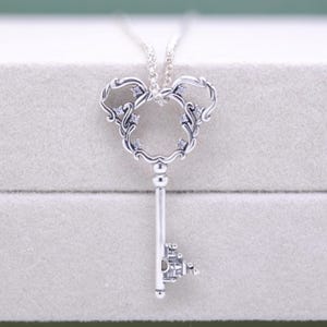 May include: A silver-colored pendant necklace featuring a key design. The key's head is shaped like a mouse with decorative swirls and small, clear gemstones. The key's stem is straight with a detailed end. The necklace chain is also silver.