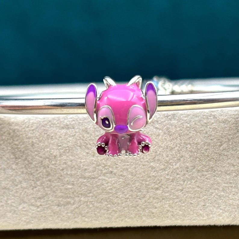 May include: A pink and purple enamel charm featuring a cartoon character with large eyes and a winking expression. The charm is made of silver and has a beaded edge.