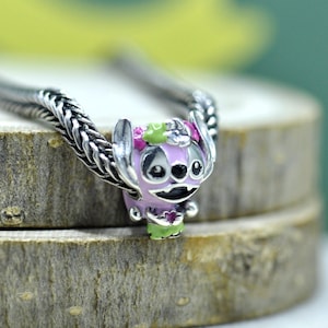 May include: A silver charm bead featuring a light purple character with large ears, a green floral crown, and a green skirt, holding a small pink heart. The charm is threaded onto a silver snake chain bracelet with a textured, braided metal link design.