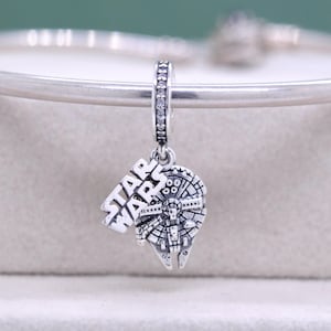 May include: A silver charm featuring the Millennium Falcon from Star Wars. The charm has the words "STAR WARS" in bold letters. The charm is suspended from a silver chain with small, clear stones.
