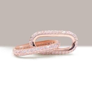 Può includere: Due maglie ovali color oro rosa, ciascuna incastonata con piccole pietre preziose trasparenti. Le maglie sono interbloccate, creando un design a catena. Le pietre preziose brillano e l'effetto complessivo è elegante e lussuoso.