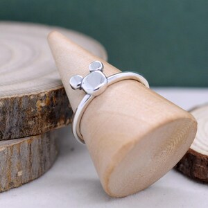 May include: A silver ring featuring a Mickey Mouse design. The ring has a round center and two smaller circles on top, resembling the iconic character's head. The ring is displayed on a wooden cone.