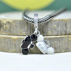 May include: A silver charm pendant featuring a black and white design of a bride and groom. The charm is suspended from a silver chain and has a decorative bail with clear stones. The bride's side has a crown and the groom's side has a top hat.