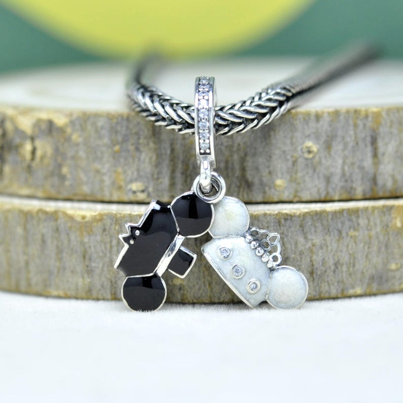 May include: A silver charm pendant featuring a black and white design of a bride and groom. The charm is suspended from a silver chain and has a decorative bail with clear stones. The bride's side has a crown and the groom's side has a top hat.
