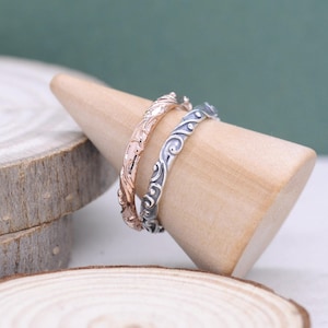 Op de afbeelding: Twee sierlijke ringen, één roségoud en één zilver, gepresenteerd op een houten kegel. De ringen hebben een gedetailleerd, wervelend patroon. De achtergrond is zachtgroen, met een houten schijf op de voorgrond.