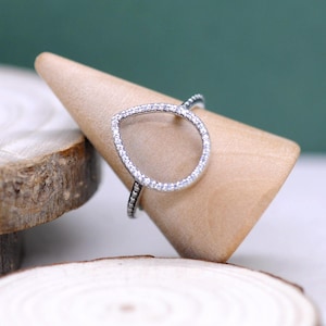 Op de afbeelding: Een zilveren ring met een druppelvormig ontwerp. De ring heeft een helder midden en is aan de buitenrand bezet met kleine, sprankelende steentjes. De ring wordt gepresenteerd op een houten ringhouder, die het elegante ontwerp benadrukt.