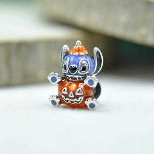 以下が含まれることがあります： 大きな耳を持つ青いキャラクターがオレンジ色のカボチャの帽子をかぶった、銀色の金属製チャームビーズ。キャラクターは、顔が彫られた大きな光沢のあるオレンジ色のカボチャを抱えています。青いエナメルのアクセントが施されています。