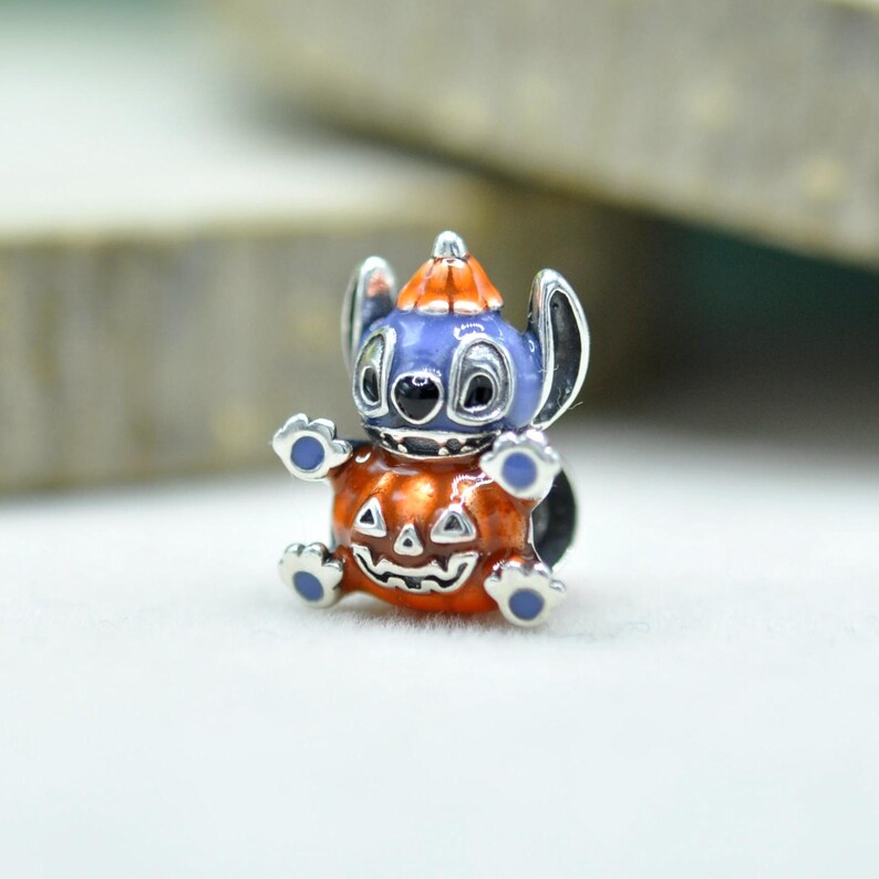 以下が含まれることがあります： 大きな耳を持つ青いキャラクターがオレンジ色のカボチャの帽子をかぶった、銀色の金属製チャームビーズ。キャラクターは、顔が彫られた大きな光沢のあるオレンジ色のカボチャを抱えています。青いエナメルのアクセントが施されています。