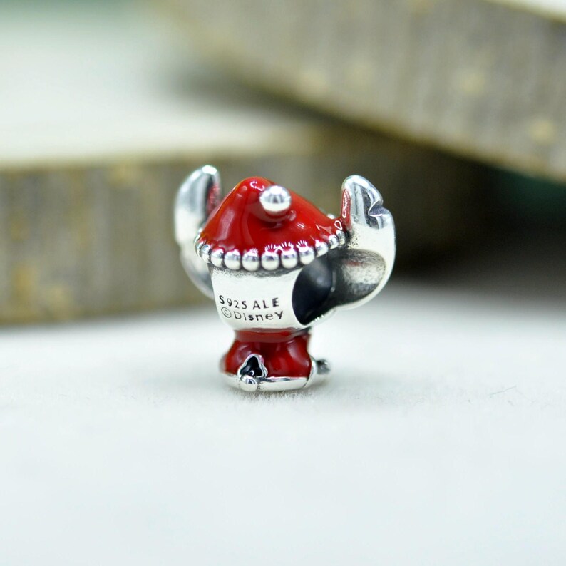 Puede incluir: Un abalorio de plata con un gorro de esmalte rojo y un borde de cuentas. La superficie met&aacute;lica est&aacute; grabada con el texto S925 ALE y Disney. El abalorio tiene una abertura central para pulsera y dos elementos laterales que parecen pinzas de cangrejo.