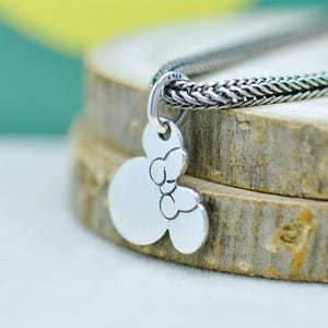 Peut inclure: Un collier pendentif en argent avec un charm en forme de Mickey Mouse. Le charm pr&eacute;sente les contours du visage et des oreilles de la c&eacute;l&egrave;bre souris. Le collier a une cha&icirc;ne tress&eacute;e. Le pendentif est pr&eacute;sent&eacute; sur une surface en bois.