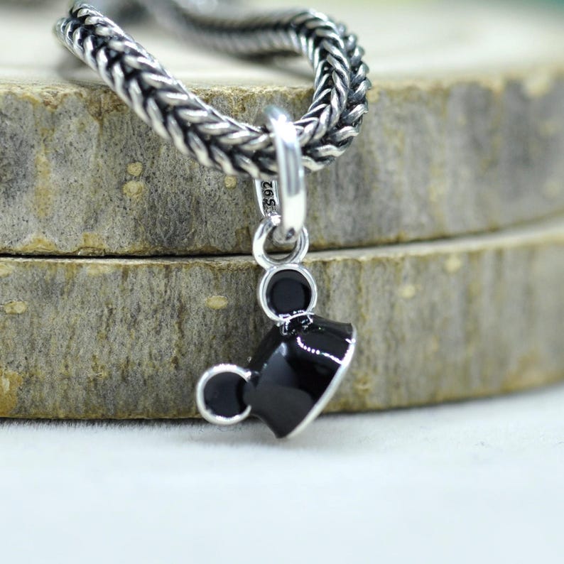 May include: A silver charm with a black graduation cap design. The charm is attached to a silver chain. The cap has a shiny black finish and a small silver accent. The charm is likely a pendant for a necklace or bracelet.