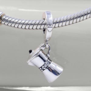 Może przedstawiać: Srebrny charms w kształcie ekspresu do kawy, z czarnym wierzchem i uchwytem. Charms jest przymocowany do srebrnej bransoletki. Charms ekspresu do kawy ma ozdobny pasek z małymi, przezroczystymi kamieniami.