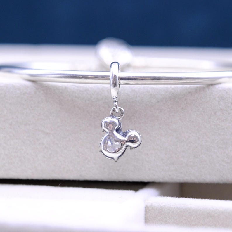 May include: Silver charm in the shape of Mickey Mouse's head, hanging from a silver bracelet.