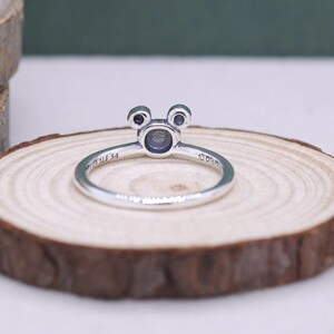 May include: A silver ring featuring a Mickey Mouse-inspired design. The ring has a circular center with two small, round accents above, resembling ears. The band is thin and polished, with the inscription "ALE 54" visible. The ring is set on a wooden surface.