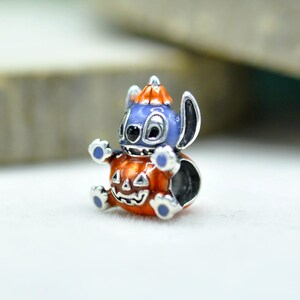 以下が含まれることがあります： 大きな耳を持つ薄紫色のキャラクターが、オレンジ色のカボチャの帽子をかぶり、笑顔の彫刻が施されたオレンジ色のハロウィーンカボチャの中に座っているシルバーチャームビーズ。足には青いエナメルのディテールが施されています。