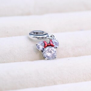 May include: A silver charm featuring a cartoon mouse with a red bow and clear crystal accents. The charm is designed to be attached to a bracelet or necklace.