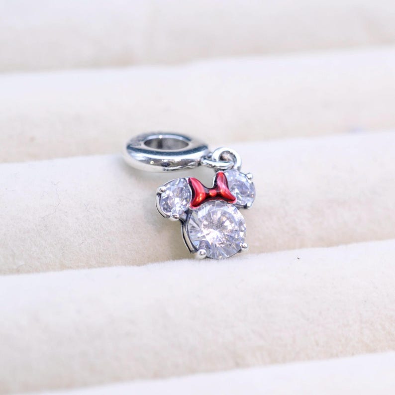 May include: A silver charm featuring a cartoon mouse with a red bow and clear crystal accents. The charm is designed to be attached to a bracelet or necklace.