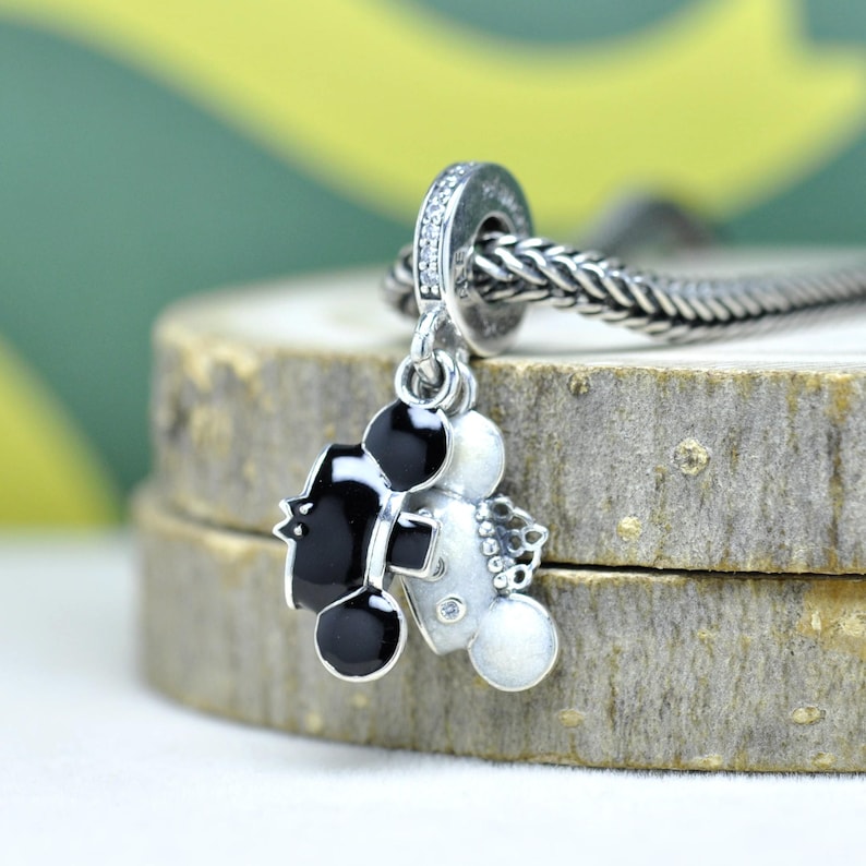 May include: A silver charm with a black and white Mickey Mouse design. The charm has a small loop with clear stones and is attached to a silver chain. The charm is on a wooden surface.