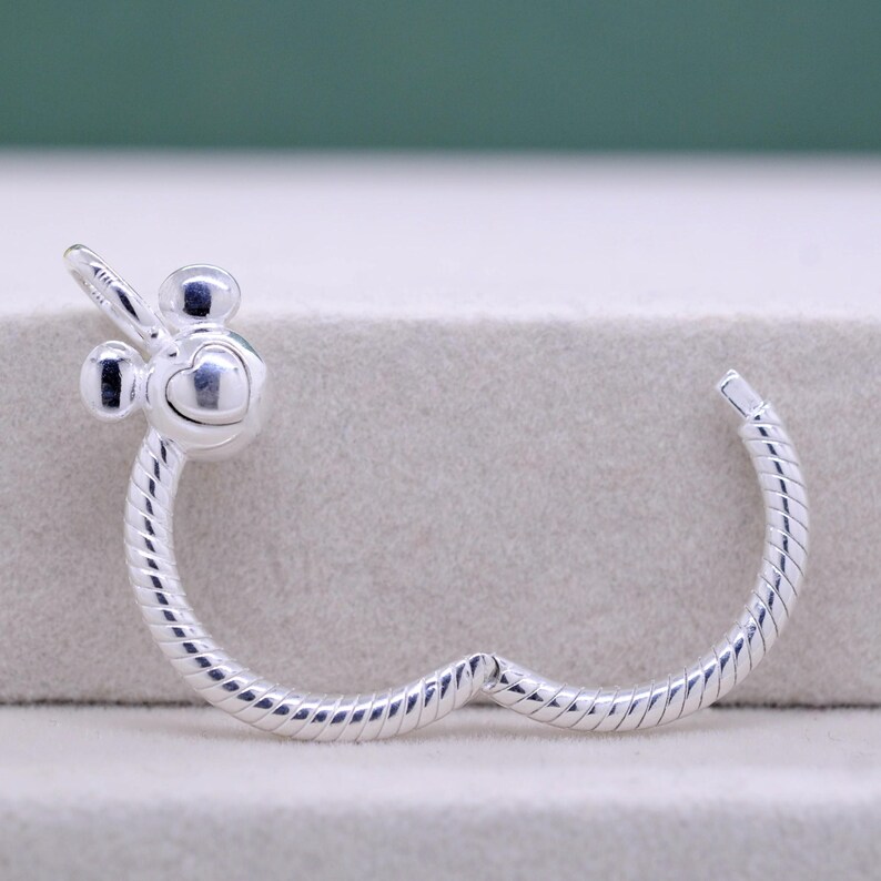 Pu&ograve; includere: Un braccialetto color argento con un design ispirato a Mickey Mouse. Il braccialetto presenta un ciondolo a forma di cuore e una fascia a spirale. Il ciondolo ha due piccole orecchie rotonde e un viso a forma di cuore. Il braccialetto &egrave; in metallo lucido.