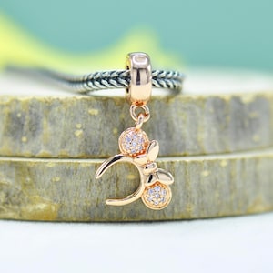 May include: Rose gold charm with a Minnie Mouse design. The charm features a round bail, a dangling bow, and two round accents, all encrusted with small, clear crystals. The charm is suspended from a silver braided chain.