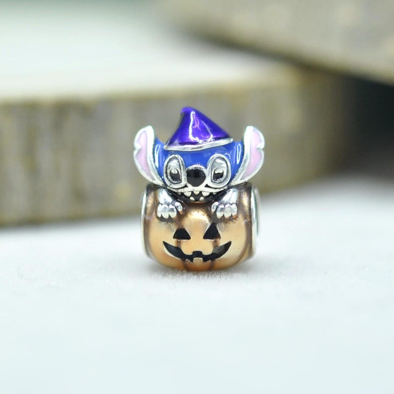 May include: A silver charm bead featuring a blue character with large ears wearing a purple pointed hat, emerging from a metallic orange pumpkin with a carved jack-o'-lantern face. The character has black eyes and a wide grin with visible teeth.