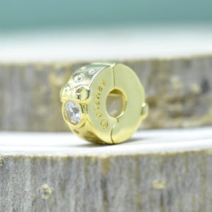 Puede incluir: Un charm dorado con una gema redonda y transparente y la palabra "Disney" grabada en el lateral. El charm es circular con una abertura de cierre. El charm est&aacute; sobre una superficie de madera.