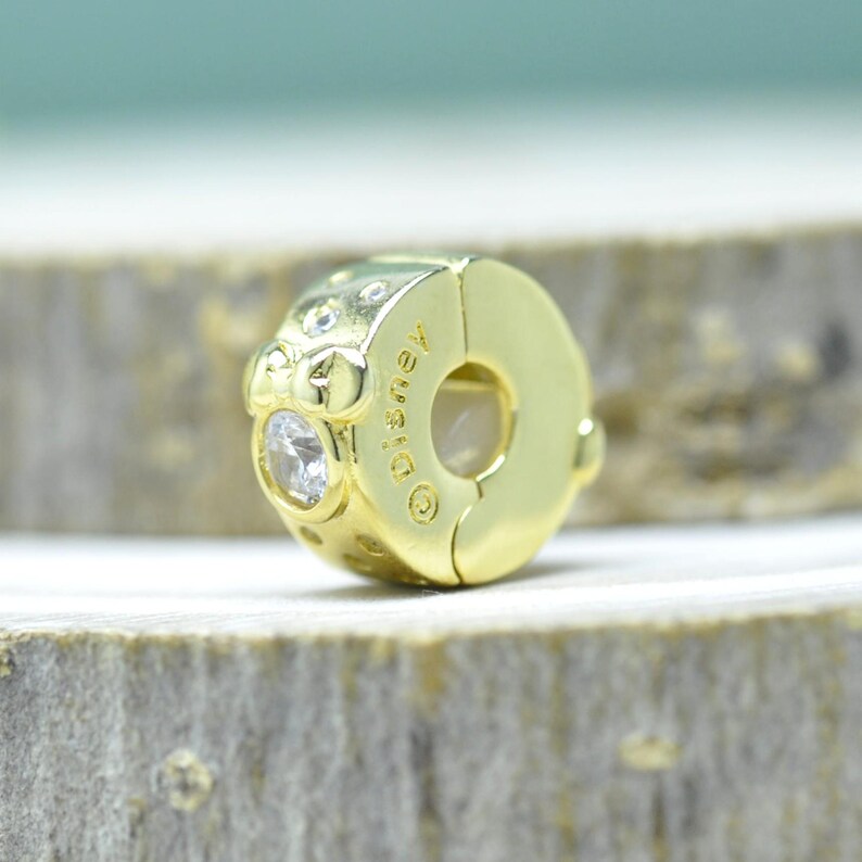 Puede incluir: Un charm dorado con una gema redonda y transparente y la palabra "Disney" grabada en el lateral. El charm es circular con una abertura de cierre. El charm est&aacute; sobre una superficie de madera.