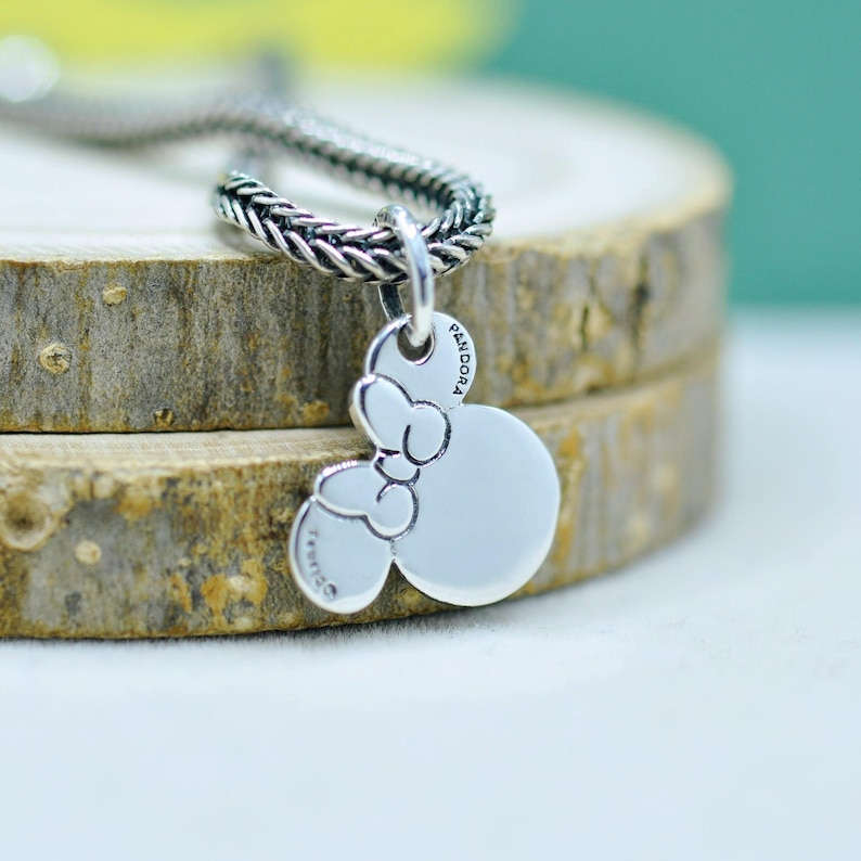 Peut inclure: Un collier avec un pendentif argent&eacute; repr&eacute;sentant Minnie Mouse. Le pendentif a un n&oelig;ud et deux formes circulaires, avec les mots "Pandora" et "&copy;Disney" grav&eacute;s. La cha&icirc;ne est de style tress&eacute;.