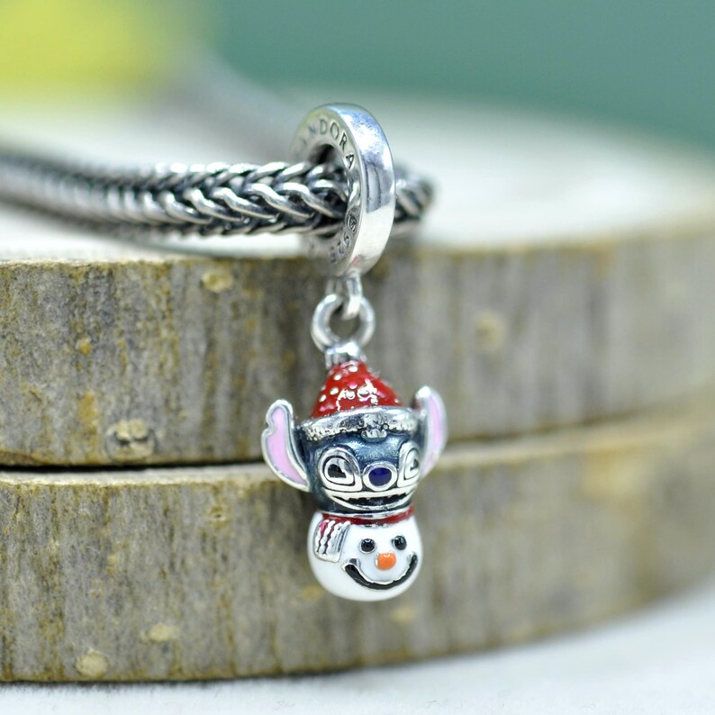 Puede incluir: Un charm de plata que muestra un personaje con orejas grandes y un gorro de Pap&aacute; Noel rojo y blanco, unido a un cuerpo de mu&ntilde;eco de nieve con nariz naranja y bufanda roja. El charm tiene grabado el nombre Pandora y cuelga de una cadena.