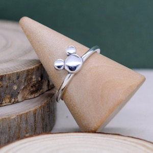 May include: A silver ring featuring a Mickey Mouse design. The ring has a polished finish and is displayed on a wooden cone. The Mickey Mouse design is composed of three circular shapes, with a larger circle in the center and two smaller circles above.