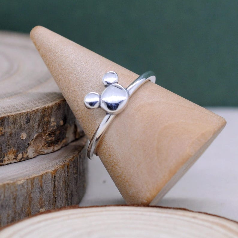 May include: A silver ring featuring a Mickey Mouse design. The ring has a polished finish and is displayed on a wooden cone. The Mickey Mouse design is composed of three circular shapes, with a larger circle in the center and two smaller circles above.