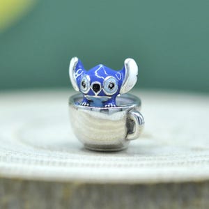May include: A silver-toned metal charm featuring a small, blue, alien-like character with large ears and white accents, sitting inside a miniature silver cup. The character has black eyes and a black nose, with a smooth, polished metallic finish.