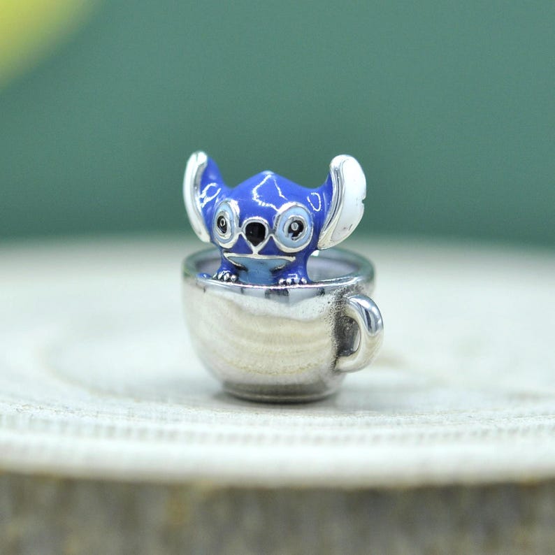 May include: A silver-toned metal charm featuring a small, blue, alien-like character with large ears and white accents, sitting inside a miniature silver cup. The character has black eyes and a black nose, with a smooth, polished metallic finish.