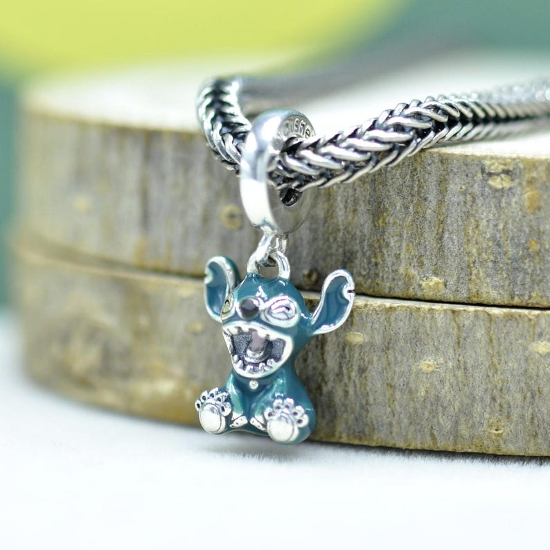 May include: A teal and silver charm featuring a cartoon character with large ears and an open mouth. The charm is suspended from a silver chain. The charm is detailed with white and black accents.