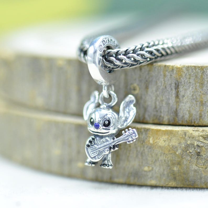 May include: A silver charm featuring the character Stitch from Disney, depicted holding a small guitar. The charm has a polished metallic finish with a small blue accent on the nose and is attached to a silver chain with a woven link pattern.