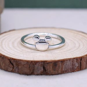 May include: A silver ring featuring a Mickey Mouse design. The ring has a circular band with a Mickey Mouse head design in the center. The ring is displayed on a wooden slice.