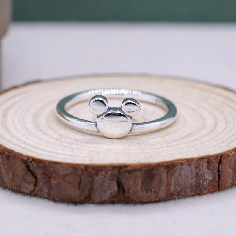 May include: A silver ring featuring a Mickey Mouse design. The ring has a circular band with a Mickey Mouse head design in the center. The ring is displayed on a wooden slice.