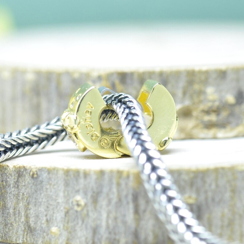 Puede incluir: Primer plano de un cierre dorado con una cadena plateada. El cierre tiene un dise&ntilde;o redondeado con una abertura con bisagras. La cadena est&aacute; hecha de eslabones plateados entrelazados. El cierre tiene la palabra "Disney" grabada.