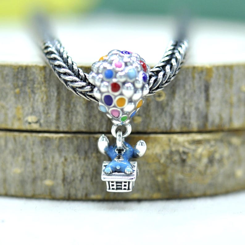 May include: A silver charm bead featuring a cluster of colorful enamel balloons attached to a small silver house with a blue character figure resting on top, all threaded onto a braided silver chain necklace with a metallic finish.