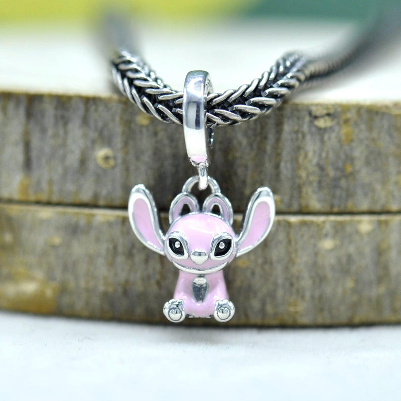 May include: A silver charm featuring a pink cartoon character with large ears and black eyes. The charm is suspended from a silver chain. The character is in a sitting position with its arms raised.