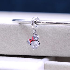 May include: Silver charm with a red bow and clear crystal accents in the shape of Mickey Mouse. The charm is attached to a silver bracelet.