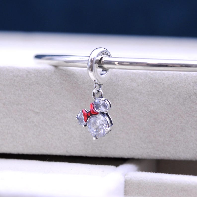 May include: Silver charm with a red bow and clear crystal accents in the shape of Mickey Mouse. The charm is attached to a silver bracelet.