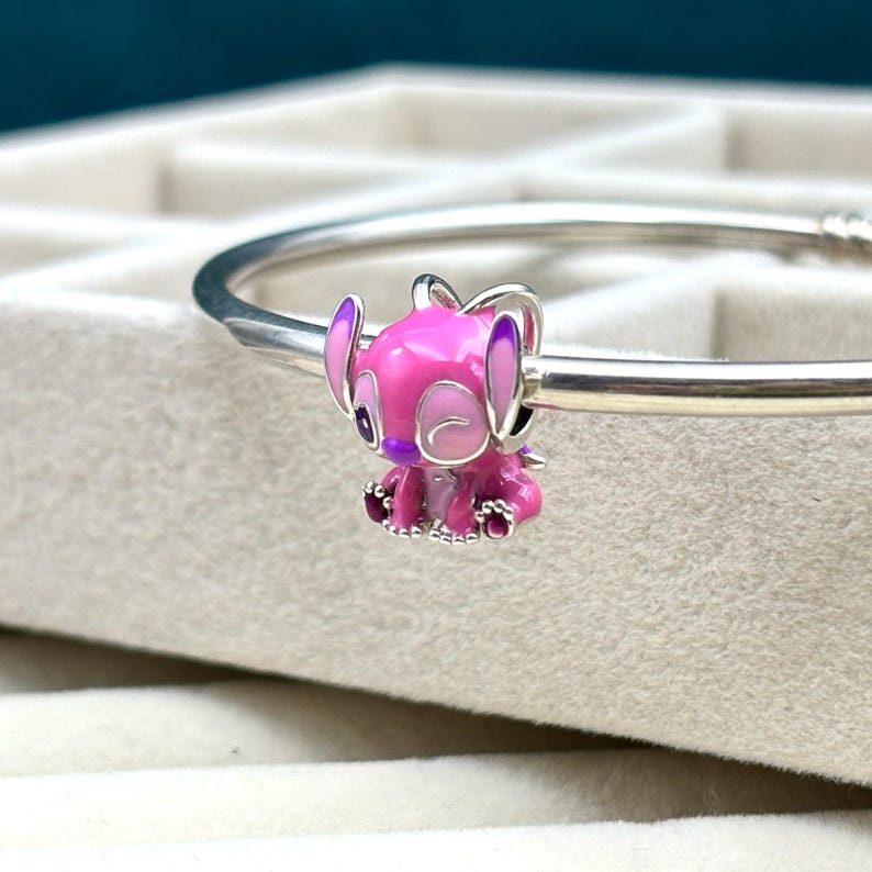 May include: A pink and purple enamel charm of a cartoon character with big eyes and ears. The charm is sitting on a silver bracelet.