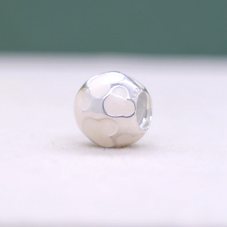 May include: A round, silver charm with a pattern of white cloud shapes. The charm has a polished finish and a small hole for threading onto a bracelet or necklace. The charm is a jewelry accessory.