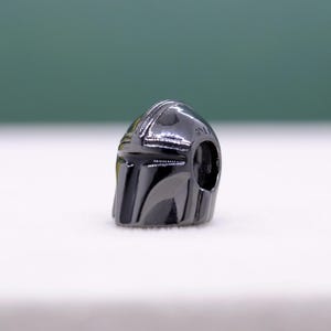 Puede incluir: Un charm met&aacute;lico gris oscuro con forma de casco. El casco tiene una superficie lisa y reflectante con detalles visibles, incluyendo una visera y elementos laterales. El charm est&aacute; dise&ntilde;ado con un agujero para ensartar.